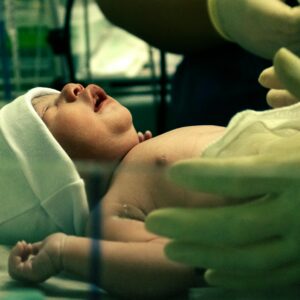 Newborn screenings are used to find medical issues. When delayed, the result may be injury or worse. Image shows a newborn on a bed with two gloved hands nearby.
