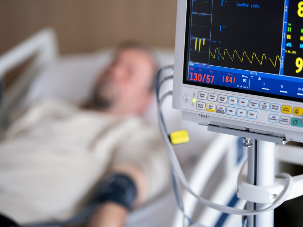 A patient in the background with a monitor in the foreground. Post-operative monitoring is crucial in ensuring that patient's benefit from their procedures while mitigating the risk of deterioration.